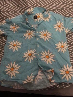 Blommig blå skjorta - Säljer en ljusblå skjorta från med stora vita blommor och orange mitt. Skjortan har korta ärmar, klassisk krage och knappar framtill. Perfekt för dig som vill sticka ut med ett färgglatt och somrigt plagg.