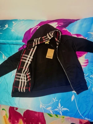 Svart hoodie från Burberry - Snygg svart hoodie från Burberry med dragkedja och klassiskt rutigt foder i huvan. Hoodien har två fickor framtill, justerbar huva med snören och diskret Burberry-logga på bröstet. Perfekt för en stilren och avslappnad look. Den är helt ny och kan även tänka mig byta mot något annat.