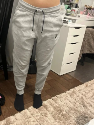 Gråa mjukisbyxor från Nike XS - Säljer ett par gråa mjukisbyxor från Nike i storlek XS. Byxorna har dragsko i midjan, muddar vid bensluten och sidofickor. Tillverkade i mjukt sweatshirtmaterial och har en liten Nike-logga på vänster ben. Perfekta för chill dagar.