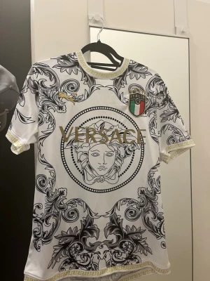 Vit Versace x Italia t-shirt - Unik vit t-shirt från Versace med svart och guld barockinspirerat mönster och stora VERSACE-bokstäver framtill. Italien-emblem på bröstet och gulddetaljer vid krage och ärmslut. Perfekt statement-plagg för dig som gillar lyxig och iögonfallande stil.