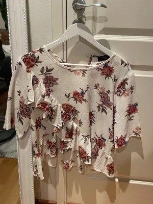 Blommig volangblus från Sisters Point strl 40 - Supersöt blus från Sisters Point i vit bas med blommigt mönster i rött, rosa och blått. Blusen har volangdetaljer framtill och på ärmarna samt rund halsringning och korta ärmar. Perfekt för dig som gillar romantisk och lekfull stil.