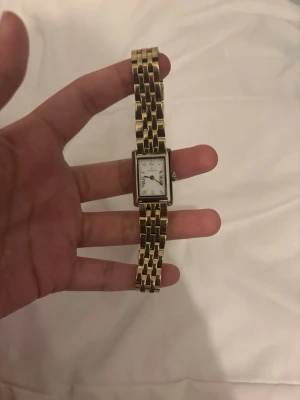 Mockberg klocka  - Stilren klocka från mockberg med rektangulärt ur och guldfärgad länk i metall. Urtavlan är vit med tydliga markeringar och klassisk design. Perfekt accessoar för dig som gillar en elegant och tidlös look. Storlek runt handleden är runt 17-18 cm. 