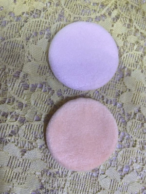 Rosa och ljuslila puderpuffar - Två runda puderpuffar i mjuk textil, en i ljusrosa och en i ljuslila. Puffarna har en slät yta på ena sidan och ett satinband på baksidan för enkel applicering av puder eller foundation. Perfekta för att få till en jämn finish på sminket. 35kr/2st/bild 