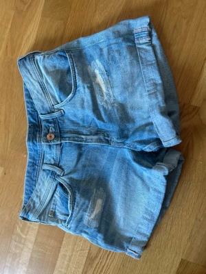 Ljusblå denimshorts med slitningar - Säljer ett par midwaist ljusblå denimshorts med uppvikta benslut och slitna detaljer framtill och baktill. Klassisk femficksmodell med knapp och dragkedja. Perfekta för varma dagar och enkel att matcha med olika toppar. Säljer då de inte kommer till användning 💕 storlek 170 men skulle säga att de passar en S