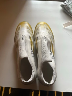 Adidas F50  - Säljer ett par Adidas F50 fotbollsskor i vitt med gula och svarta detaljer. Skorna har en strömlinjeformad design utan snörning och är tillverkade i ett lätt syntetmaterial för maximal fart på planen. 