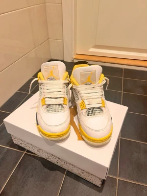 Air Jordan 4 Retro 'Tour Yellow' - Snygga Air Jordan 4 Retro sneakers i vitt läder med gula och svarta detaljer. Skorna har meshpaneler, gula snörhål och en ikonisk Jumpman-logga på plösen. Sulan är vit med gula och svarta inslag. Perfekta för dig som vill sticka ut med klassisk streetwearstil.