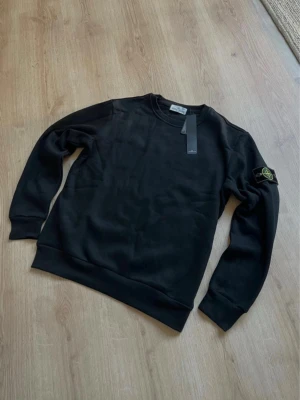 Stone Island sweatshirt - Svart sweatshirt från Stone Island med klassisk rund hals och ribbade muddar. Tröjan har den ikoniska logotypen på vänster ärm och är tillverkad i mjukt bomullsmaterial. Perfekt för en clean och stilren look.