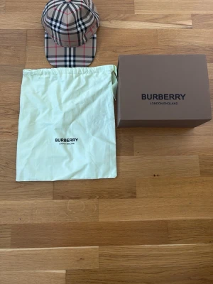Burberry beige rutig keps - Säljer en klassisk Burberry keps i beige med det ikoniska rutmönstret i svart, vitt och rött. Kepsen har böjd skärm och justerbar rem bak. Tillverkad i bomull och levereras med originalpåse och box. Perfekt accessoar för en trendig streetwear-look.
