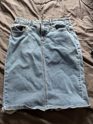 Blå jeanskjol med slits - Säljer en klassisk blå jeanskjol i rak modell med fyra fickor och slits bak. Kjolen har bälteshällor och stängs med knapp och dragkedja framtill. Perfekt till vardags för en avslappnad look.