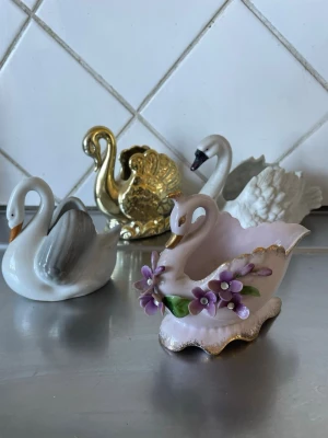 Porslinsprydnader svanar mixade färger - Fyra dekorativa svanfigurer i porslin och keramik, varav en är guldfärgad, en vit med grå vingar, en vit och en ljusrosa med lila blommor och guldkant. Perfekta som prydnad till hyllan eller fönsterbrädan.ca från 10- 16 cm långa 