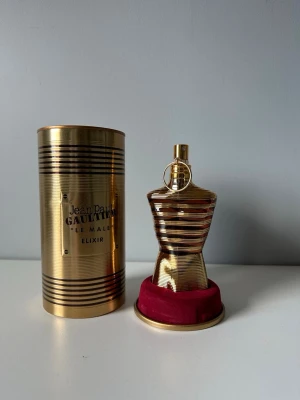 Jean Paul Gaultier Le Male Elixir - Jean Paul Gaultier Le Male Elixir parfym i en ikonisk guldfärgad flaska formad som en manlig torso. Flaskan har metallisk finish och levereras med en matchande guld- och svart randig metalltub samt en röd sammetsbas. Perfekt för dig som gillar lyxig design.