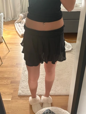 Svart volangkjol i chiffong - Säljer en svart kortkjol med volangdetaljer och flowy känsla. Kjolen är gjord i ett tunt, lätt material som ger en luftig look. Perfekt för att styla med crop tops eller stickade tröjor. Supersnygg och enkel att matcha!