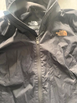 Svart vindjacka från The North Face - Snygg svart vindjacka från The North Face med dragkedja framtill och huva. Jackan har logga broderad på bröstet och är tillverkad i ett lätt, vindtätt material. Perfekt för blåsiga dagar och enkel att packa ner i väskan.