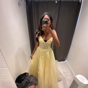 Ljusgul långklänning med spets och glitter - Drömmig ljusgul långklänning med smala axelband, v-ringning och glittrande tyll. Överdelen har vackra spetsdetaljer och broderier som ger en sagolik känsla. Kjolen är voluminös och skimrar snyggt i ljuset – perfekt för bal eller student! Orginalpris 6000kr