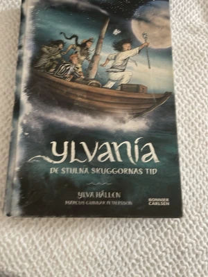 Ylvania: De stulna skuggornas tid - En spännande fantasybok av Ylva Hällen med illustrationer av Marcus Gunnar Pettersson. Omslaget är färgstarkt med blå och gröna toner och visar fyra barn i en båt på ett stormigt hav under natthimmel. Boken är inbunden och rikt illustrerad, perfekt för dig som gillar äventyr.