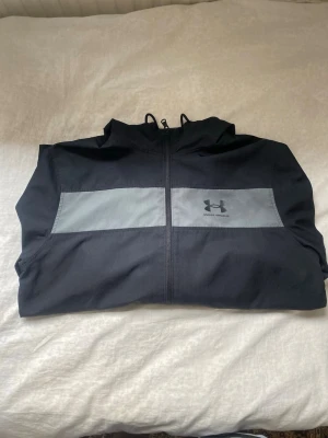 Svart vindjacka från Under Armour - Snygg svart vindjacka från Under Armour med grått band över bröstet och logga på vänster sida. Jackan har dragkedja framtill, huva och meshfoder för extra komfort. Perfekt för blåsiga dagar och sportiga outfits.