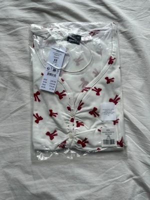 Vit pyjamaströja med röda rosetter - Supersöt vit pyjamaströja från Gina Tricot med röda rosetter över hela tyget. Tröjan har v-ringning och knappar framtill. Perfekt för mysiga kvällar hemma. Tillverkad i mjukt material för extra komfort.