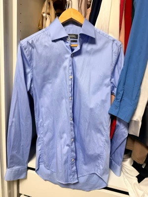 Ljusblå slim fit skjorta Dressmann - Säljer en ljusblå skjorta från Dressmann i slim fit-modell. Skjortan är tillverkad i stretchig bomull, har klassisk krage, långa ärmar och knäppning framtill. Perfekt för dig som gillar en stilren och enkel look.