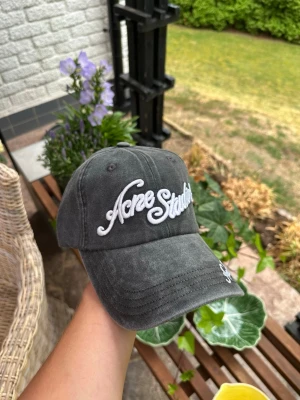 Mörkgrå keps från Acne Studios - Säljer en mörkgrå keps från Acne Studios med broderad vit logga framtill och på skärmen. Kepsen har klassisk böjd skärm och justerbar rem bak. Materialet är bomull med en tvättad vintage-look. Perfekt för en avslappnad streetwear-stil.