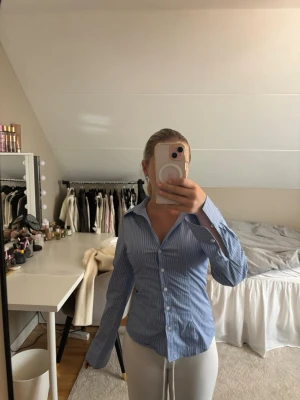 Blåvit randig skjorta med knappar - Säljer en klassisk blåvit randig skjorta med knappar framtill och krage. Skjortan har lång ärm och en figurnära passform som ger en snygg siluett. Perfekt till jeans eller kjol för en stilren look.