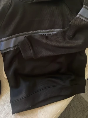 Svart hoodie med reflexdetalj - Snygg svart hoodie med huva och en bred reflexliknande rand över bröstet och ena ärmen. Hoodien har ribbade muddar vid ärmslut och nederkant. Perfekt för dig som gillar en stilren look med lite edge.