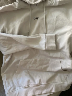 Vit hoodie från Off-White - Säljer en vit hoodie från Off-White med liten 'OFF' text på bröstet och coola svarta penseldrag på ryggen. Hoodien har huva med dragsko och stor magficka framtill. Perfekt för dig som gillar streetwear och vill sticka ut.