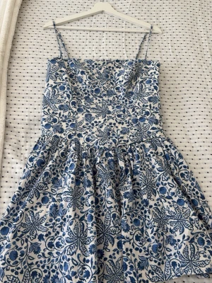 Blåvit blommig kortklänning - Söt blåvit klänning med smala axelband och blommigt mönster. Klänningen har en utställd kjol och är perfekt för varma dagar. Tillverkad i ett lätt material som känns skönt mot huden.