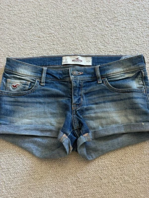 Blå jeansshorts från Hollister - Snygga blå jeansshorts från Hollister med klassisk femficksdesign och uppvikta benslut. Shortsen har slitna detaljer och en liten logga på fickan fram. Perfekta för varma dagar och har en avslappnad passform.💓