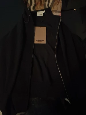Svart vindjacka från Burberry - Snygg svart vindjacka från Burberry med klassiskt rutigt foder i huvan och dragkedja framtill. Jackan har en modern passform och är perfekt för dig som vill ha en stilren look med exklusiv känsla.
