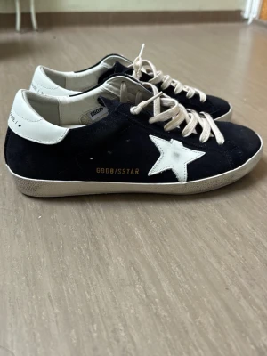 Mörkblå Golden Goose Superstar  - Säljer ett par mörkblåa Golden Goose Superstar sneakers med vit stjärndetalj på sidan. Skorna är tillverkade i mocka och läder.                                       Endast använda någon gång för att testa men de passade tyvärr inte. Ej ÄK ___ TA.       Det tillkommer låda och dustbag till. Skriv för mer info!