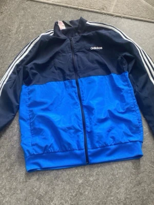 Adidas blue and black colorblock zip-up jacket - Säljer en snygg vindjacka från Adidas i blå och marinblå färg. Jackan har dragkedja framtill, klassiska vita ränder längs ärmarna och Adidas-logga på bröstet. Tillverkad i lätt och tåligt syntetmaterial, perfekt för en sportig look.