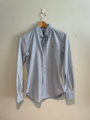 Ljusblå skjorta från Ralph Lauren - Klassisk ljusblå skjorta från Ralph Lauren med button-down krage och broderad logga på bröstet. Skjortan har lång ärm, vita knappar och är tillverkad i bomull. Perfekt för en clean och stilren look.