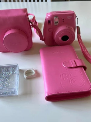 Rosa Instax Mini digitalkamera - Säljer en rosa Instax Mini digitalkamera med matchande fodral, handledsrem och tillbehör. Kameran har en kompakt och rundad design i plast, perfekt för att ta direktbilder. Medföljer även ett rosa fodral och ett glitteretui för bilder.