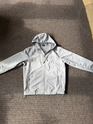 Ljusgrå vindjacka från Under Armour - Snygg ljusgrå vindjacka från Under Armour med huva och dragkedja framtill. Jackan har två sidofickor med dragkedja, elastiska muddar vid ärmsluten och justerbar dragsko nedtill. Perfekt för blåsiga dagar och sportiga outfits.