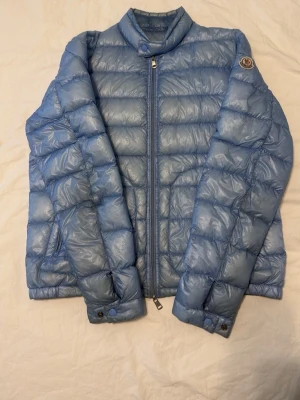 Ljusblå dunjacka från Moncler - Ljusblå dunjacka från Moncler med quiltad design och hög krage. Jackan har dragkedja framtill, sidofickor med dragkedja och Moncler-logga på ärmen. Perfekt för kalla vinterdagar och riktigt snygg till streetwear-stilen. Inga hål eller liknande men materialets färg har små förändringar som kan synas i vissa ljus 