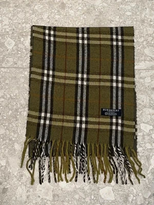 Olivgrön rutig halsduk Burberry - Snygg olivgrön halsduk från Burberry med klassiskt rutmönster i svart, vitt och brunt. Halsduken har fransar i ändarna och är tillverkad i mjuk ull. Perfekt accessoar för att lyfta din outfit och hålla dig varm.