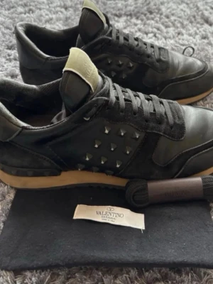 Svarta Valentino sneakers med nitar - Snygga svarta sneakers från Valentino med coola nitar på sidorna och beige detaljer på plösen och sulan. Skorna har en klassisk låg silhuett, snörning och är tillverkade i en mix av läder och mocka. Perfekta för dig som vill sticka ut med en exklusiv touch.