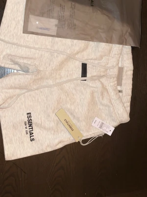 Essentials ljusbeige shorts med snörning - Säljer ett par ljusbeige shorts från Essentials med snörning i midjan och tryckt logga på benet. Shortsen har en relaxed passform och är tillverkade i mjuk bomull, perfekta för chill dagar. Kommer med originaltaggar och påse.