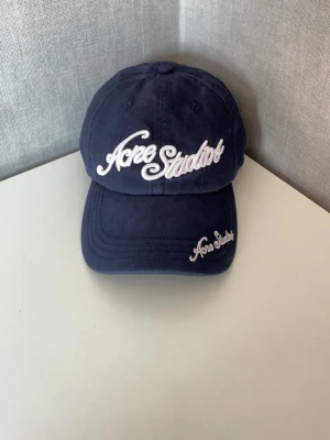 Acne Studios Keps i mörkblå - Snygg mörkblå keps från Acne Studios med vit broderad logga framtill och på skärmen. Klassisk böjd skärm och justerbar baksida. Tillverkad i mjukt bomullstyg som ger en avslappnad och trendig look.