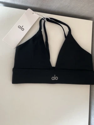 ALO sport BH - Snygg svart bh från Alo med triangelkupor och smala axelband. Bh:n har en bred resår under bysten med Alo-logga framtill. Tillverkad i ett mjukt och stretchigt material som ger en sportig look.