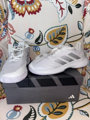 Vita Adidas sneakers med meshdetaljer - Snygga vita sneakers från Adidas med klassiska tre ränder i silver på sidan. Ovandelen är i meshmaterial för bra andningsförmåga och skon har vita snören samt en robust vit sula. Perfekta för en sportig och stilren look.