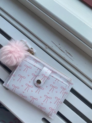 Vit plånbok med rosa rosett-mönster - Supersöt vit plånbok med rosa rosetter och små hjärtan. Plånboken har ett fack med tryckknapp, dragkedja upptill och en fluffig ljusrosa pompom som detalj. Perfekt för kort och mynt, och ger en gullig touch till din stil.