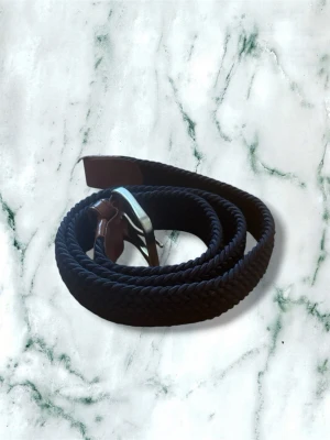Mörkblått flätat bälte med silverdetalj - Snyggt mörkblått flätat bälte med silverfärgat spänne och bruna skinn-detaljer vid ändarna. Perfekt för att ge din outfit en stilren touch. Bältet är flexibelt och passar till många olika looks.