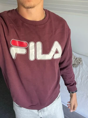 Vinröd sweatshirt från FILA - Står storlek L men passar storlek S. Säljer en vinröd sweatshirt från FILA med stort logotryck i vitt och rött på bröstet. Tröjan har rund halsringning, ribbade muddar vid ärmslut och nederkant samt är tillverkad i mjukt material. Perfekt för en avslappnad och sportig stil.