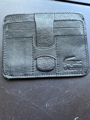 Svart korthållare från Lacoste - Snygg svart korthållare från Lacoste i läder med präglad logga framtill. Flera kortfack och en praktisk rem mitt på. Baksidan har ett fack i textil för ID eller busskort. Perfekt för dig som vill ha stilren och smidig förvaring av kort.