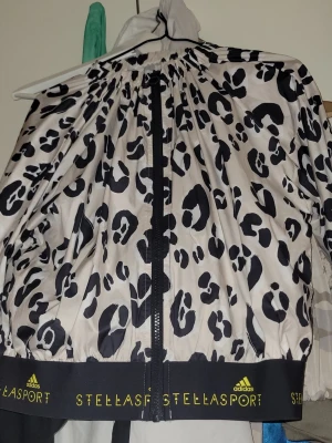 Leopardmönstrad vindjacka Adidas - Snygg vindjacka från Adidas Stellasport med svartvitt leopardmönster. Jackan har dragkedja framtill, hög krage och elastisk mudd med Stellasport-logga i gult. Perfekt för dig som vill sticka ut med sportig stil.