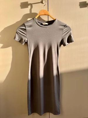Grå kortärmad t-shirtklänning - En enkel och stilren grå t-shirtklänning med rund halsringning och korta ärmar. Klänningen har en rak passform och är tillverkad i ett mjukt, stretchigt material som känns bekvämt mot huden. Perfekt för en avslappnad look.
