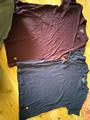 Två basic t-shirts i vinröd och marinblå - Säljer två stilrena t-shirts i vinröd och marinblå med liten gul broderad logga vid nederkanten. Båda har rund hals och korta ärmar. Perfekta till jeans eller shorts och gjorda i mjukt bomullstyg för skön känsla hela dagen.
