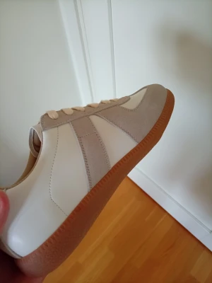 Maison margiela sneakers - Snygga sneakers i vitt och beige med ovandel i läder och mocka. Skorna har en klassisk låg siluett och en ljusbrun gummisula med mönstrad undersida för bra grepp. Perfekta för en clean och stilren look.