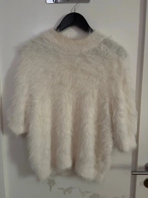 Fluffig vit stickad tröja - Supermjuk och fluffig vit stickad tröja med lös passform och rund hals. Tröjan har ett lurvigt material som ger en cool och mysig look, perfekt för kyliga dagar. Ärmarna är något kortare och modellen är oversized. Storlek: S